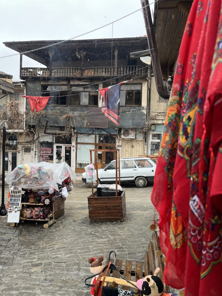 Brostensbelagt gade med en hvid bil, trækasser, træbænk og rødt tørklæde og et træhus i baggrunden