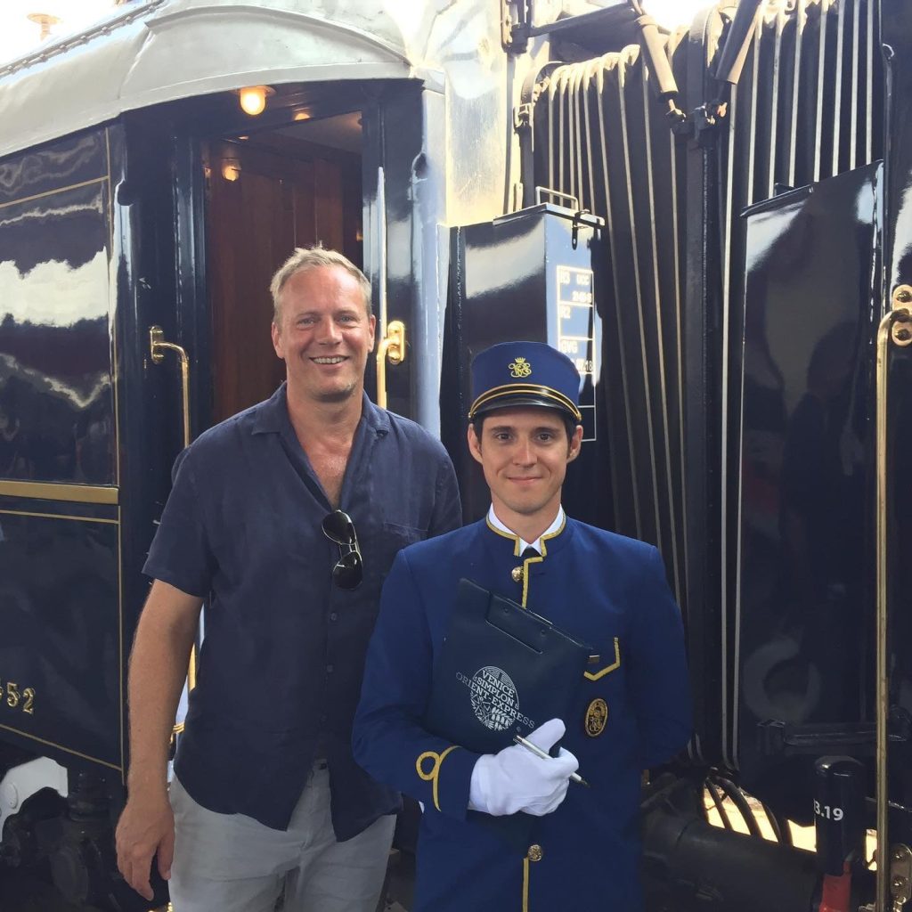 En mand i afslappet tøj og en mand i blå uniform, der står foran et sort tog af ældre dato