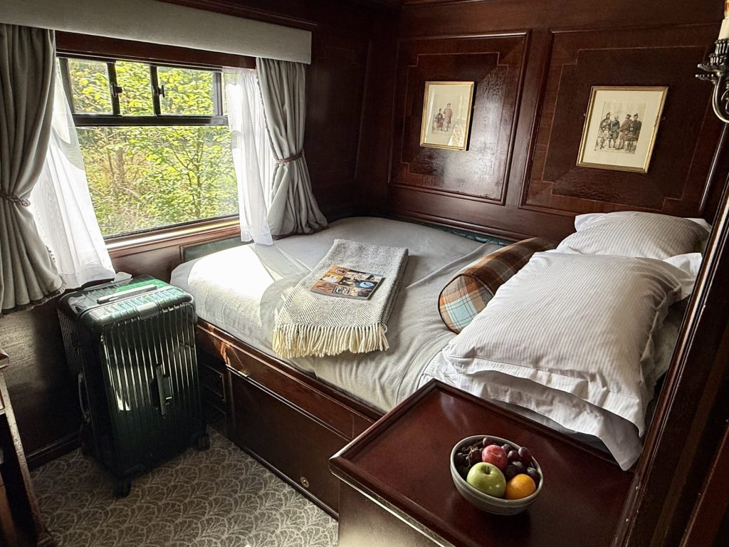 En togkupé i mørkt træ med et vindue og en dobbeltseng med hvidt sengetøj og en skål med frugt
