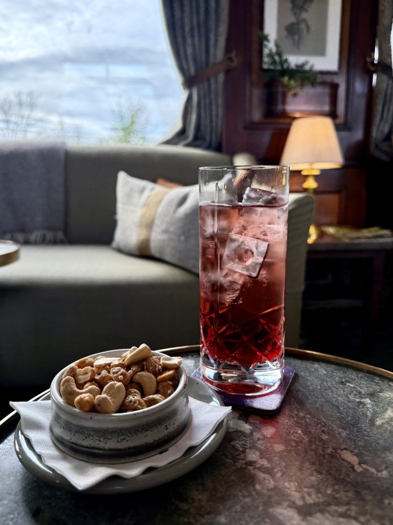 Et rundt bord med en drink med isterninger i et højt glas med en skål nødder ved siden af