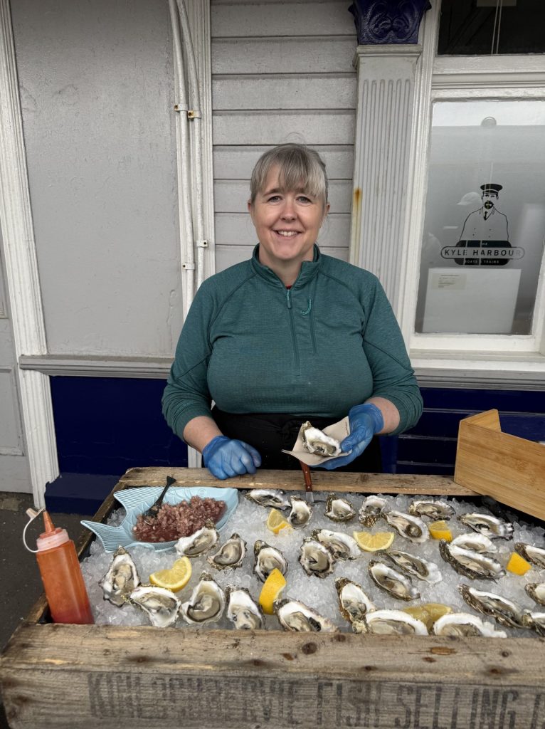 Kvinde der står med en østers i hånden mens der ligger flere åbne østers foran hende på et underlag af is