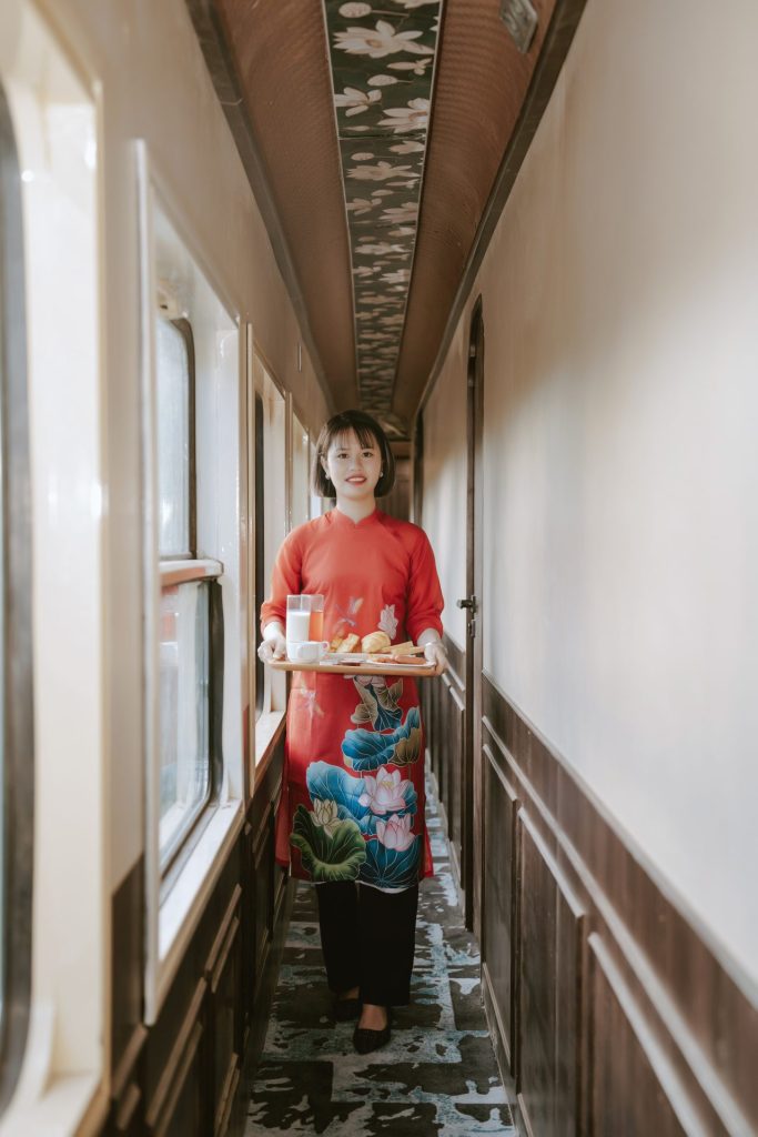 En kvinde i en rød kjole med en bakke med morgenmad stående i en mal gang i en togvogn